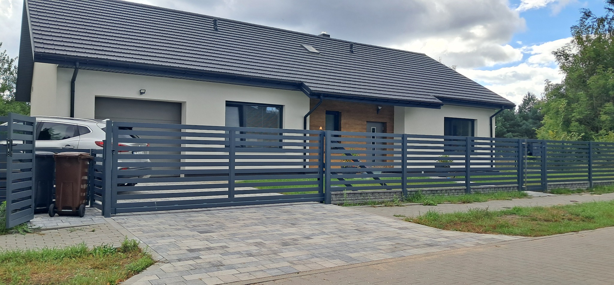 Nowoczesny dom jednorodzinny z szarą, poziomą, metalową bramą i ogrodzeniem. Na podjeździe kostka brukowa w odcieniach szarości. Widoczny fragment samochodu i kosze na śmieci.
