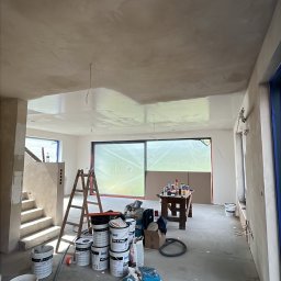 Piotr Dziedzic - Wnętrze w trakcie prac wykończeniowych: świeżo położona gładź na suficie, drabina, farby, narzędzia. Okno zabezpieczone folią malarską. Widok na schody.