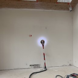 Piotr Dziedzic - Szlifowanie ściany szlifierką z odkurzaczem w jasnym wnętrzu z oknami dachowymi. Detal z prac wykończeniowych w Węgierskiej Górce.