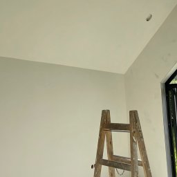 Piotr Dziedzic - Świeżo pomalowane wnętrze z drewnianą drabiną, widoczne gniazdko elektryczne wystające z sufitu i fragment okna. Jasne, naturalne światło.