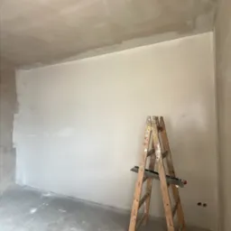 Pokój w trakcie prac wykończeniowych: ściany po szpachlowaniu, drabina z narzędziami, surowy beton na podłodze. Widoczne gniazdka elektryczne.