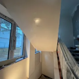 Klatka schodowa w trakcie prac wykończeniowych: gładź na ścianach i suficie, okno z widokiem na budynek. Schody zabezpieczone folią ochronną.