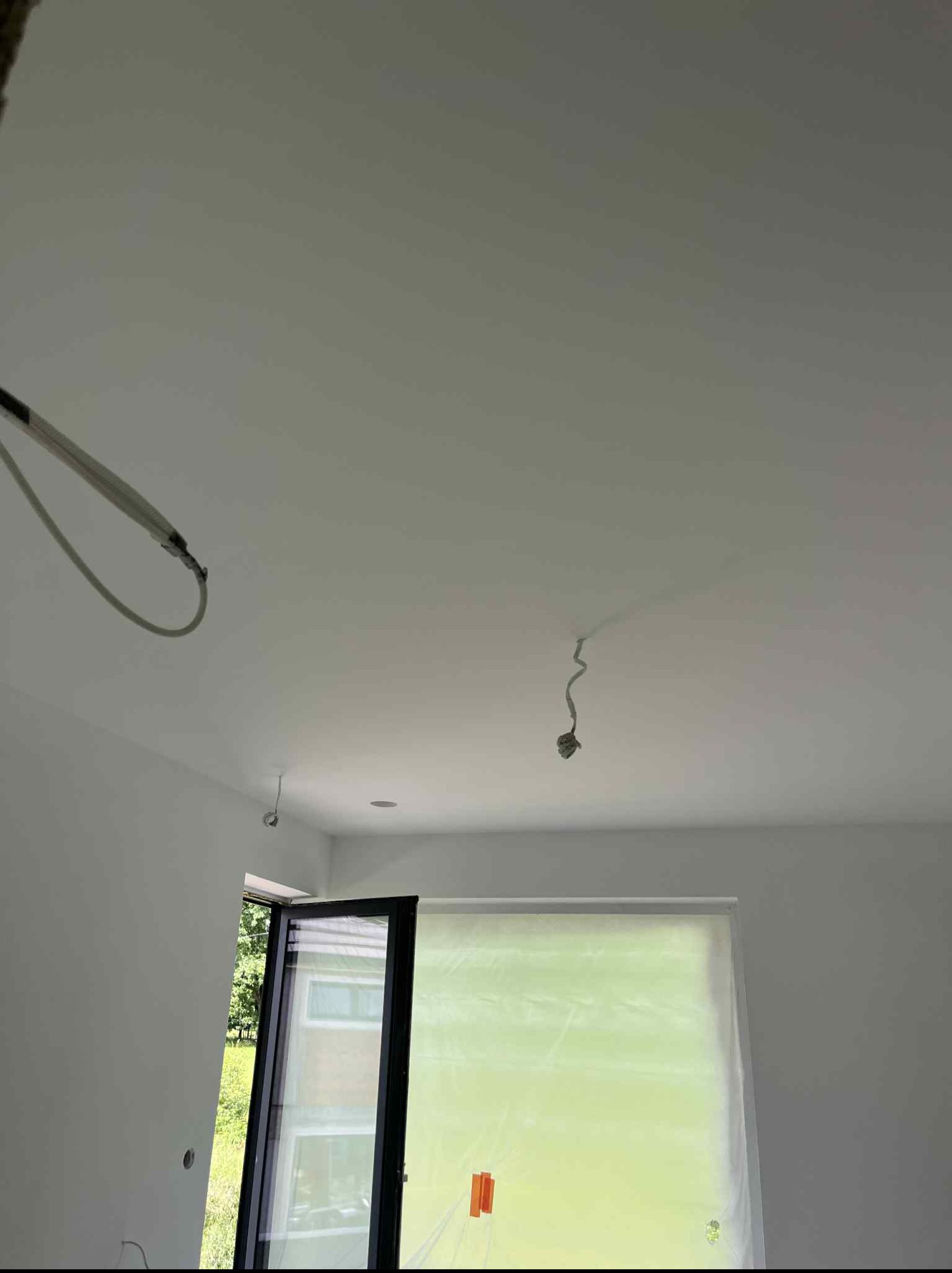 Wnętrze po malowaniu: Świeżo pomalowany sufit i ściany na biało. Widoczne kable oświetleniowe i okno zabezpieczone folią malarską. Minimalistyczny design.
