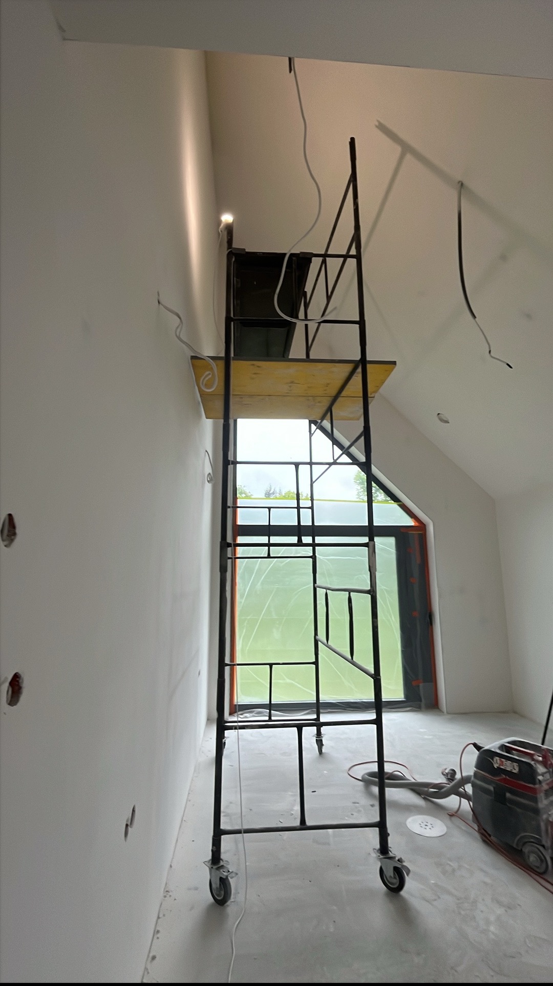 Wnętrze w trakcie wykończenia: ściany po gładzi, rusztowanie na kółkach, okno zabezpieczone folią, szlifierka do gładzi.