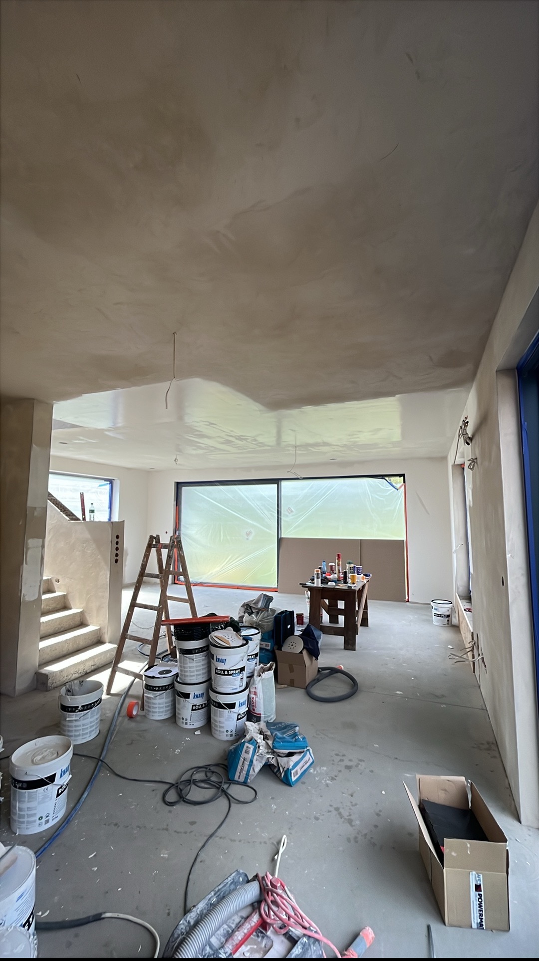 Wnętrze w trakcie prac wykończeniowych: świeżo położona gładź na suficie, drabina, farby, narzędzia. Okno zabezpieczone folią malarską. Widok na schody.