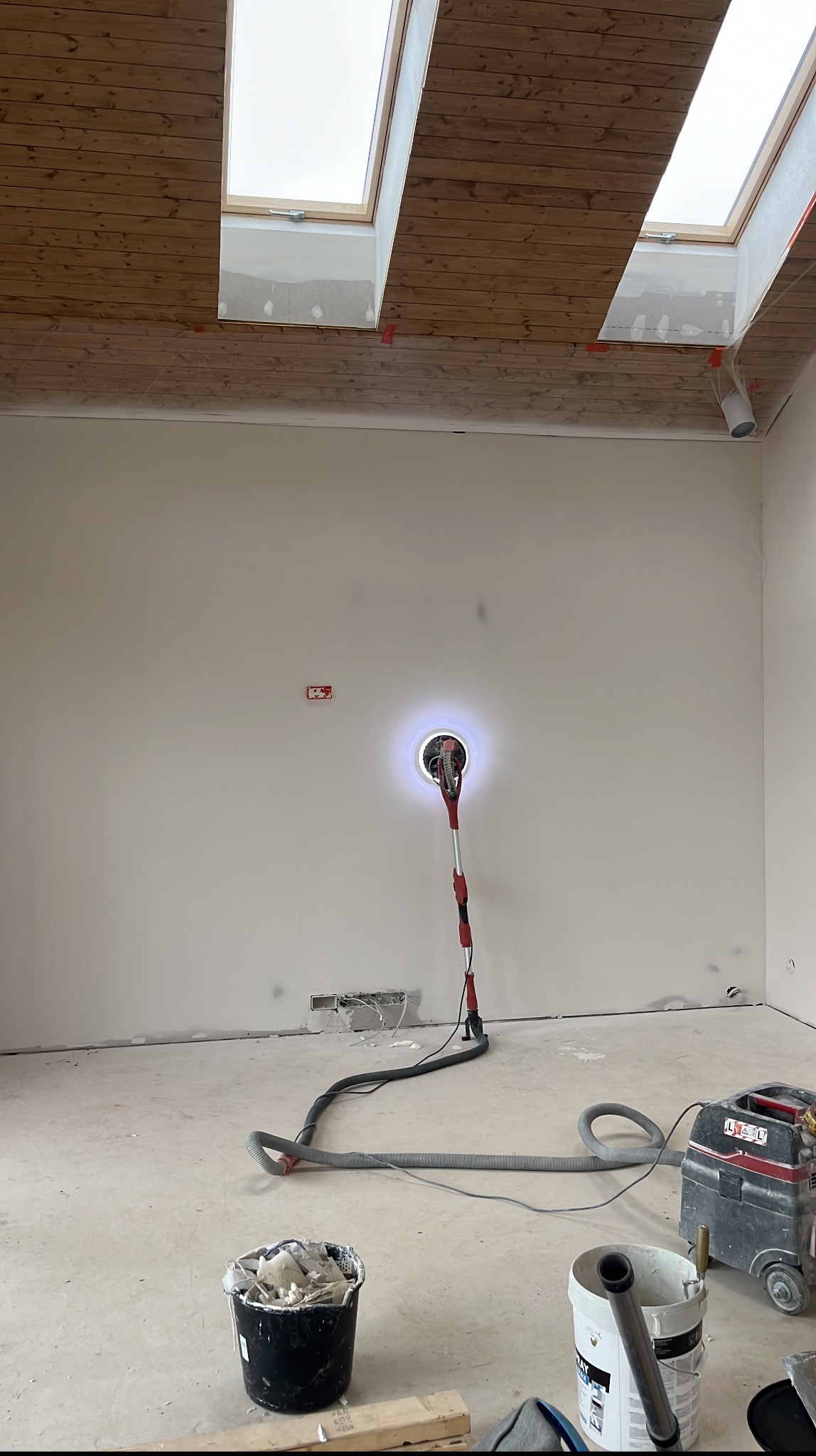 Szlifowanie ściany szlifierką z odkurzaczem w jasnym wnętrzu z oknami dachowymi. Detal z prac wykończeniowych w Węgierskiej Górce.