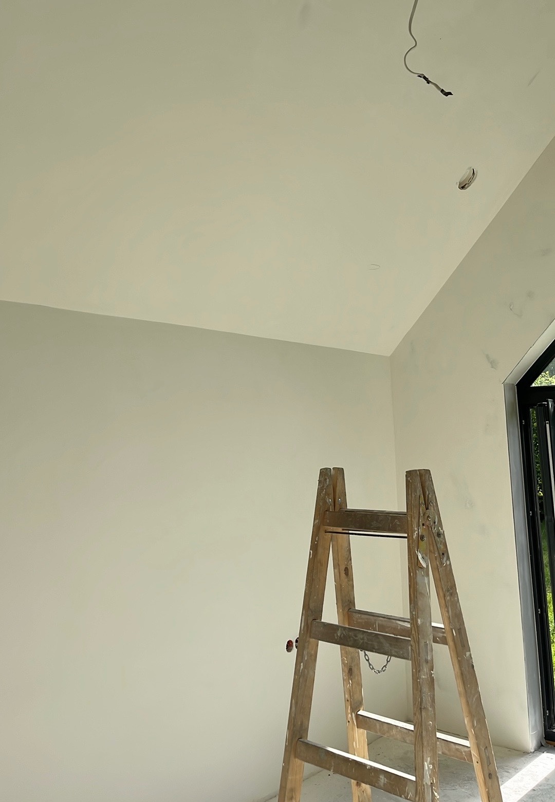 Świeżo pomalowane wnętrze z drewnianą drabiną, widoczne gniazdko elektryczne wystające z sufitu i fragment okna. Jasne, naturalne światło.