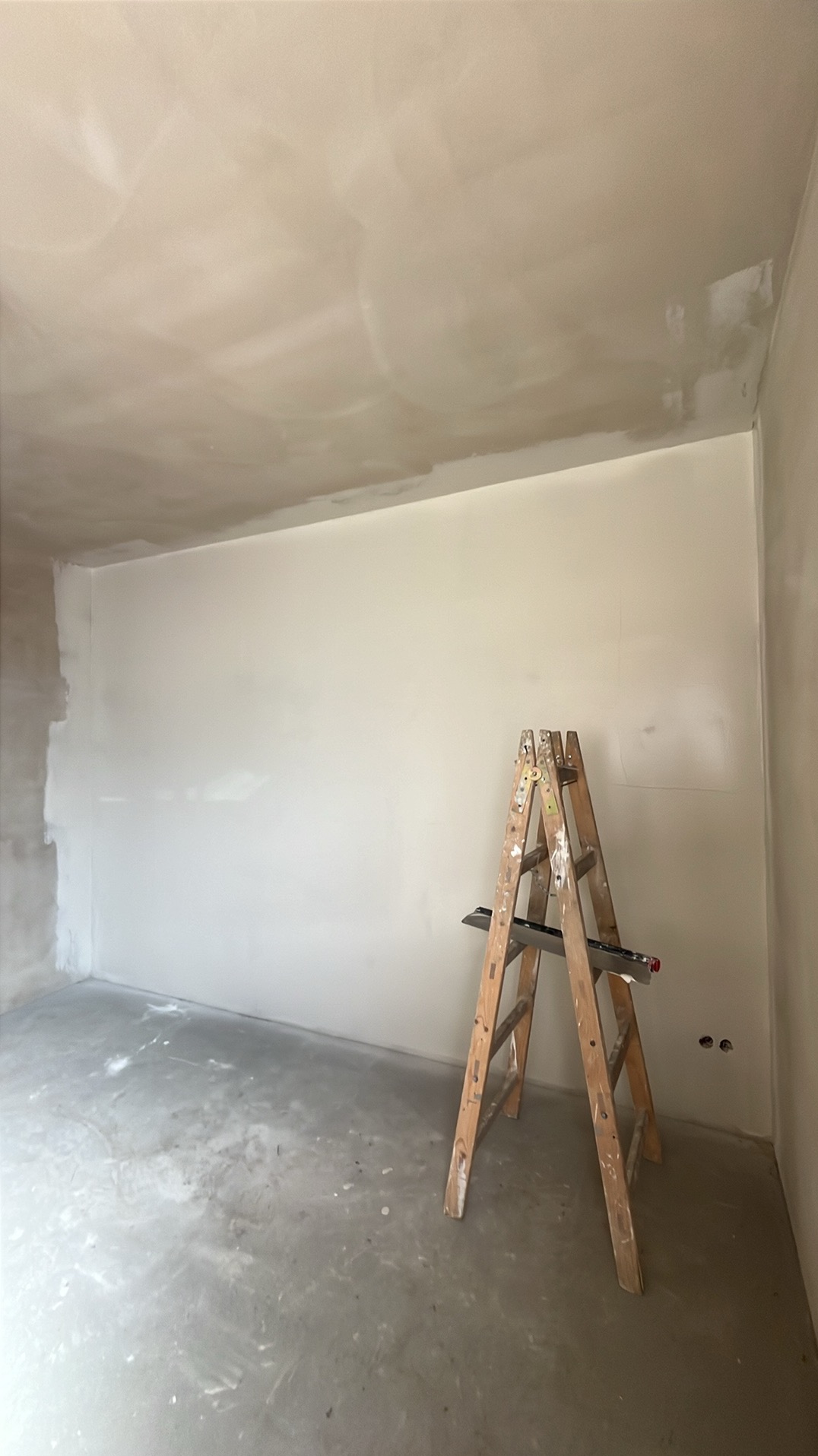 Pokój w trakcie prac wykończeniowych: ściany po szpachlowaniu, drabina z narzędziami, surowy beton na podłodze. Widoczne gniazdka elektryczne.