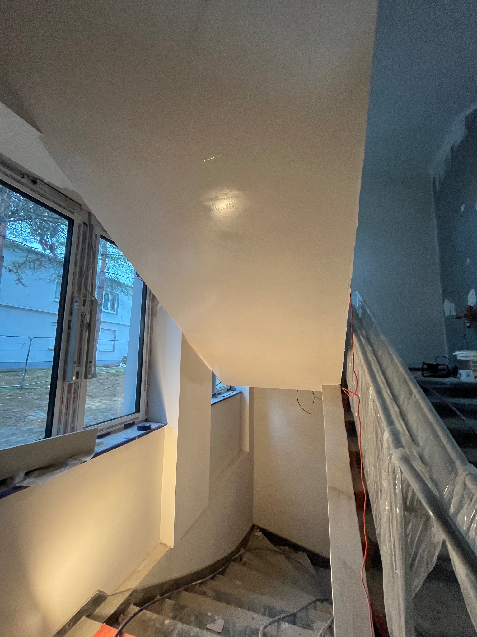Klatka schodowa w trakcie prac wykończeniowych: gładź na ścianach i suficie, okno z widokiem na budynek. Schody zabezpieczone folią ochronną.