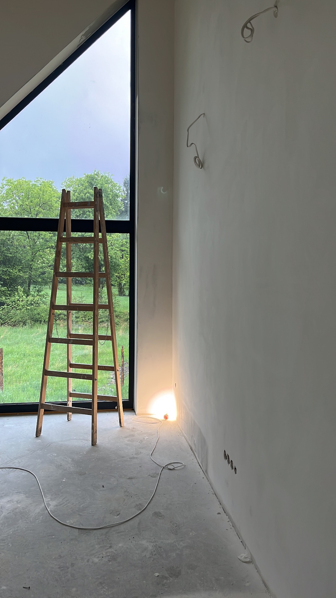 Wysoka drabina oparta o okno w surowym wnętrzu. Ściany przygotowane do malowania, widoczne przewody elektryczne. Podłoga betonowa, surowy stan wykończenia.