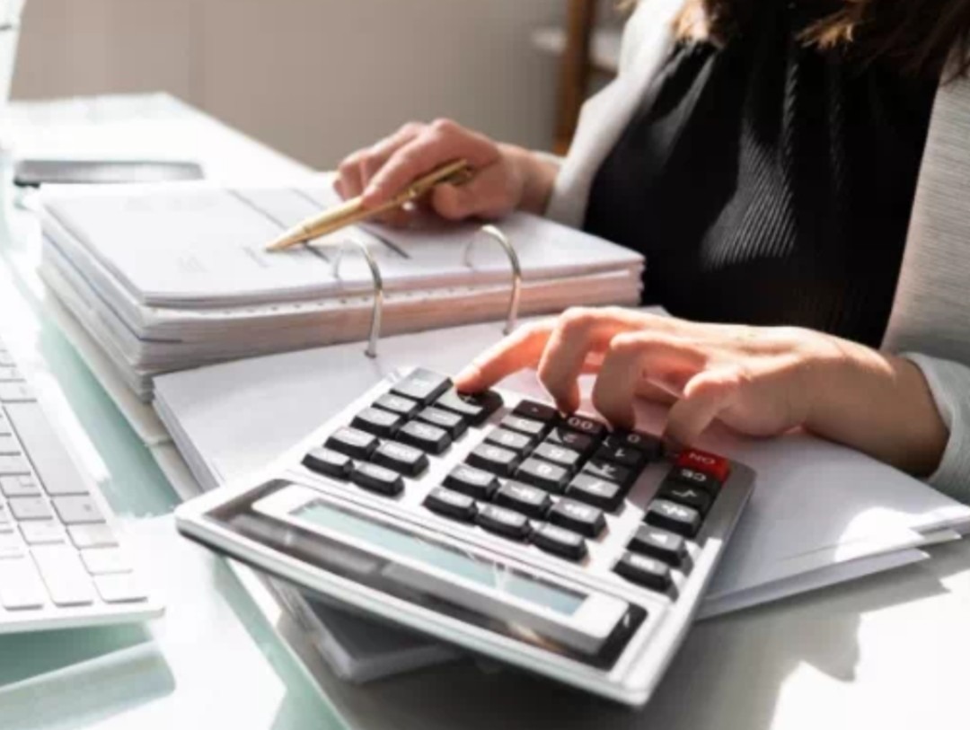 Kobieta liczy na kalkulatorze przy biurku z dokumentami i złotym długopisem. Praca biurowa, finanse, księgowość.