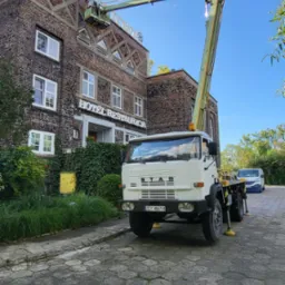 Podnośnik koszowy STAR przy budynku hotelu z cegły, kosz z osobą przy napisie na dachu. Widoczny fragment chodnika z kostki i zaparkowany bus.