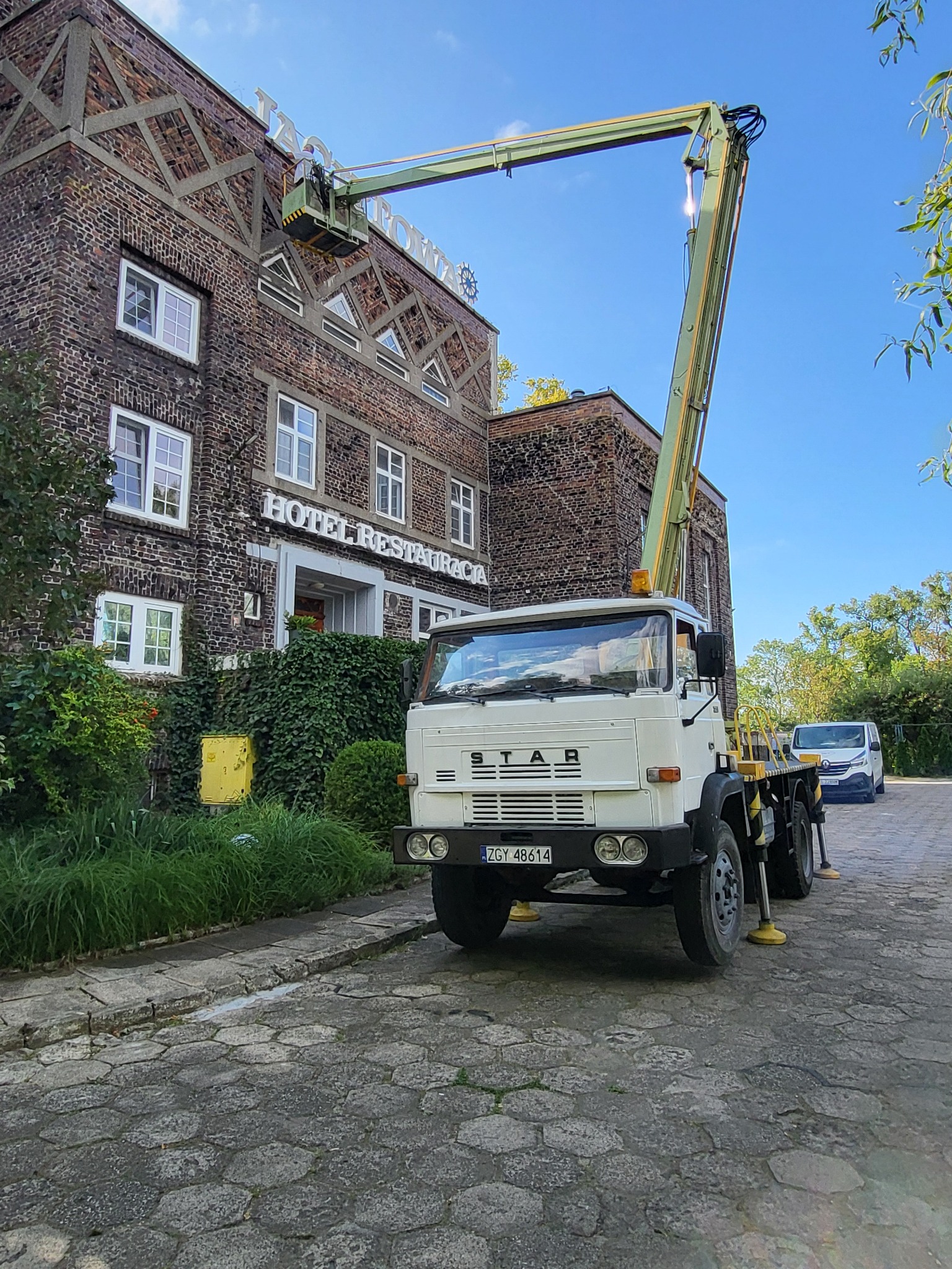 Podnośnik koszowy STAR przy budynku hotelu z cegły, kosz z osobą przy napisie na dachu. Widoczny fragment chodnika z kostki i zaparkowany bus.