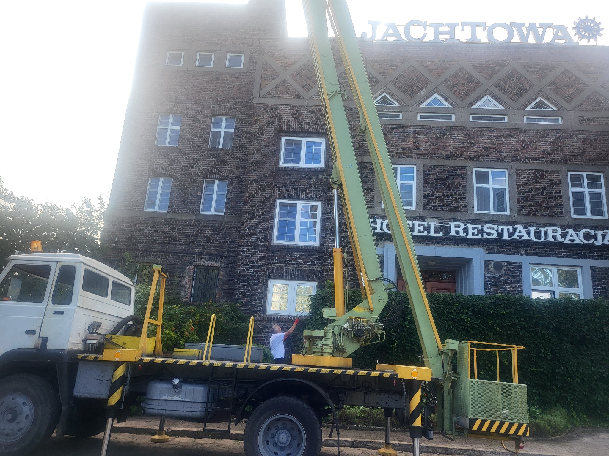 Żółto-zielony podnośnik koszowy na tle budynku z cegły, pracownik z czerwoną rękawicą przy oknie. Widoczny fragment hotelu/restauracji.