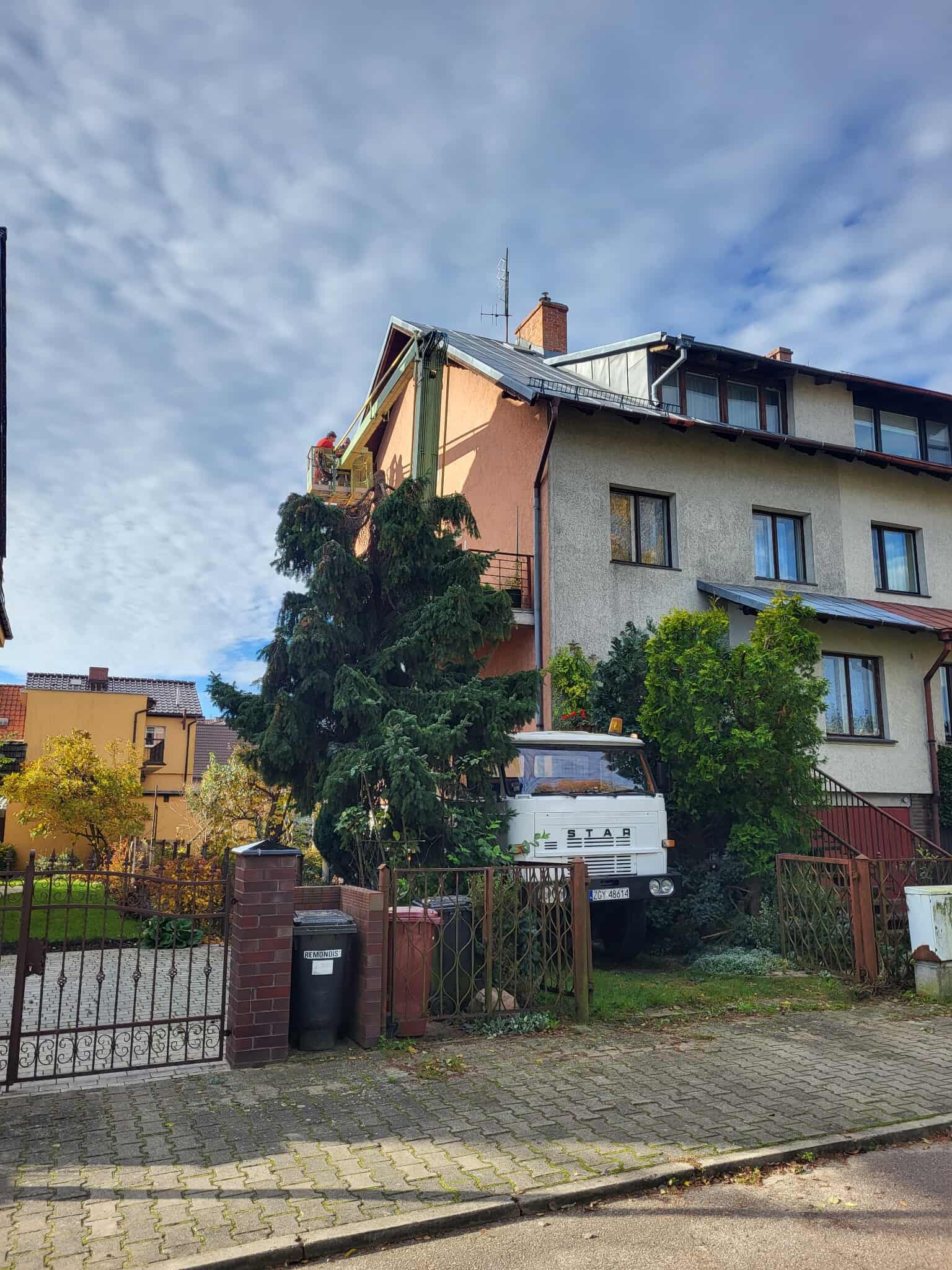 Pracownik w czerwonym ubraniu na podnośniku koszowym przy budynku, wykonuje prace na wysokości. Na pierwszym planie drzewo i zaparkowany biały samochód STAR.