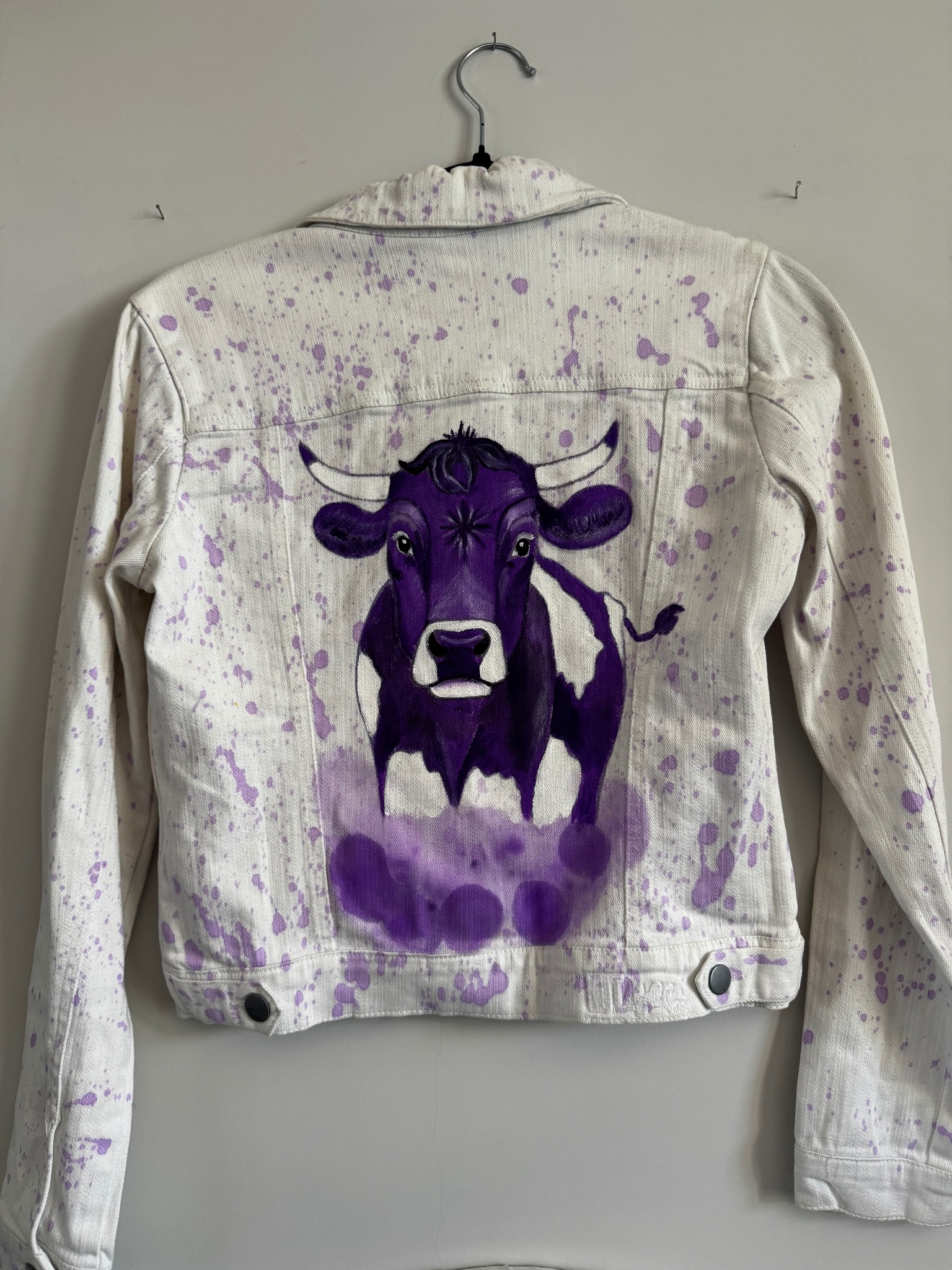 Biała kurtka jeansowa z ręcznie malowanym, fioletowym motywem krowy na plecach, wisząca na wieszaku na tle szarej ściany. Widoczne fioletowe plamy farby.