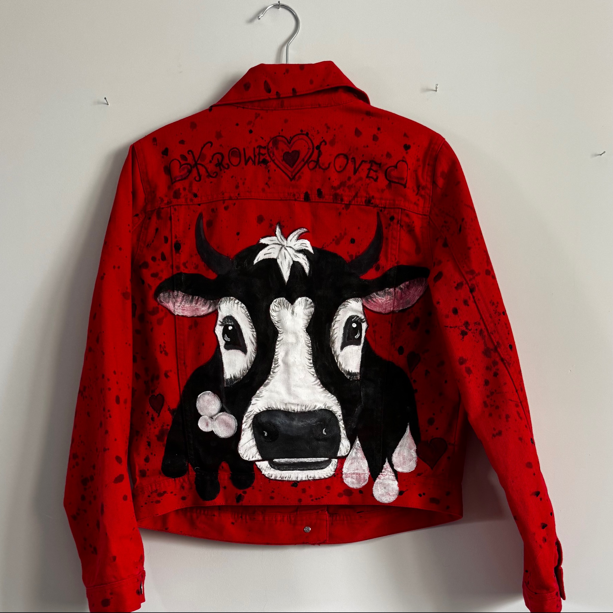 Czerwona kurtka jeansowa z ręcznie malowaną krową, wisząca na wieszaku. Widoczne detale malunku i napisy 'Krowe Love'.