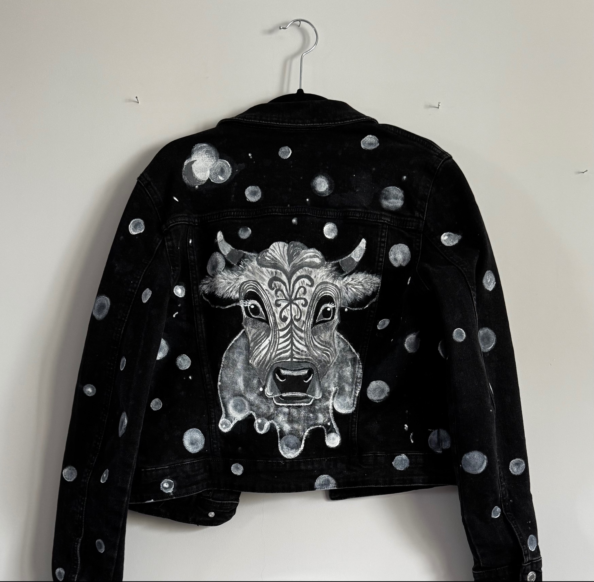 Czarna kurtka jeansowa z ręcznie malowanym motywem krowy i białymi kropkami, wisząca na wieszaku na tle jasnej ściany z widocznymi haczykami.