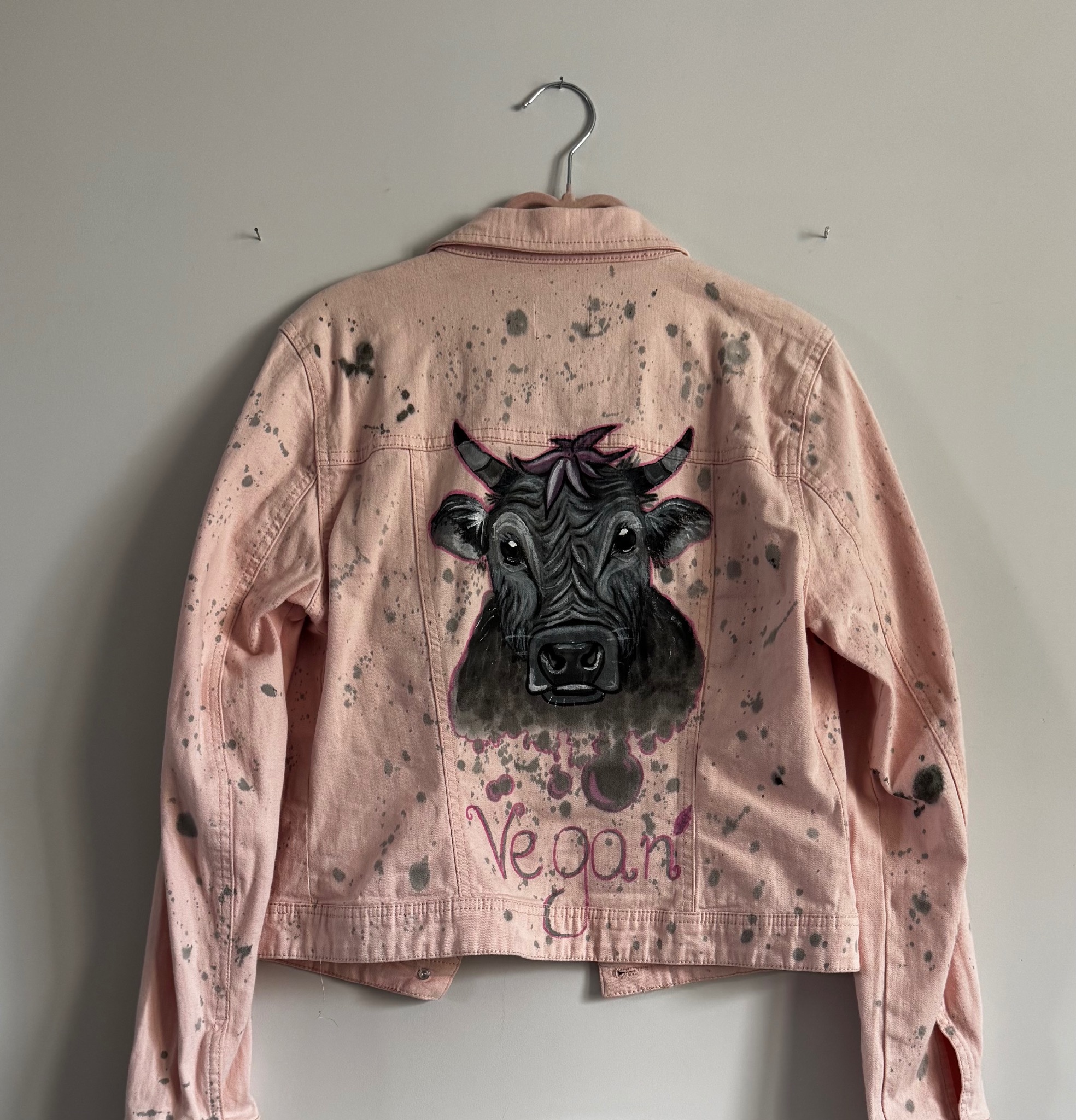 Różowa kurtka jeansowa z ręcznie malowanym motywem krowy i napisem 'Vegan!', wisząca na wieszaku na tle białej ściany. Widoczne plamy farby i unikalny design.