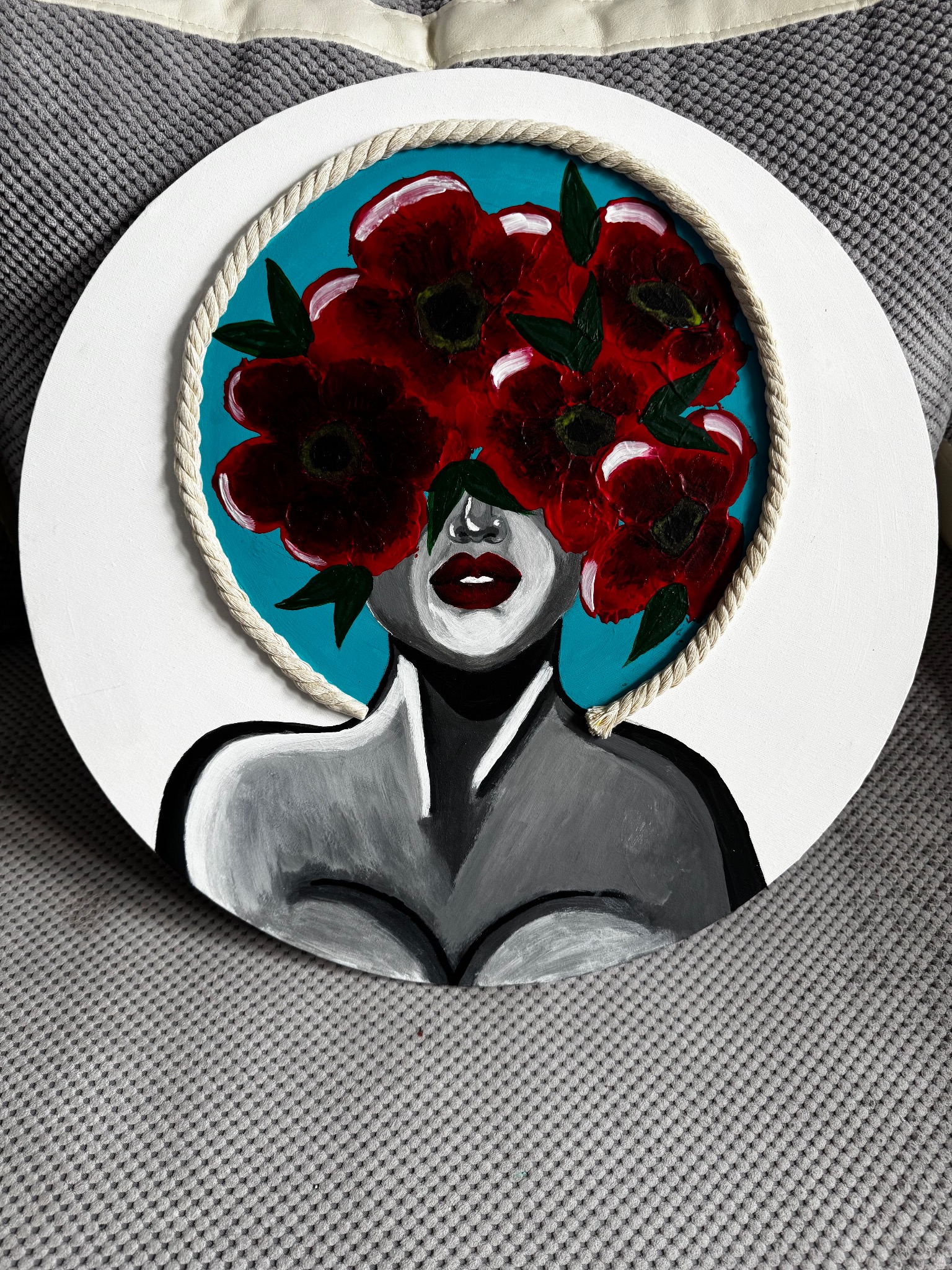Okrągły obraz na płótnie: abstrakcyjna postać kobiety z czerwonymi kwiatami zamiast włosów, turkusowe tło, całość otoczona sznurem i białą ramą.