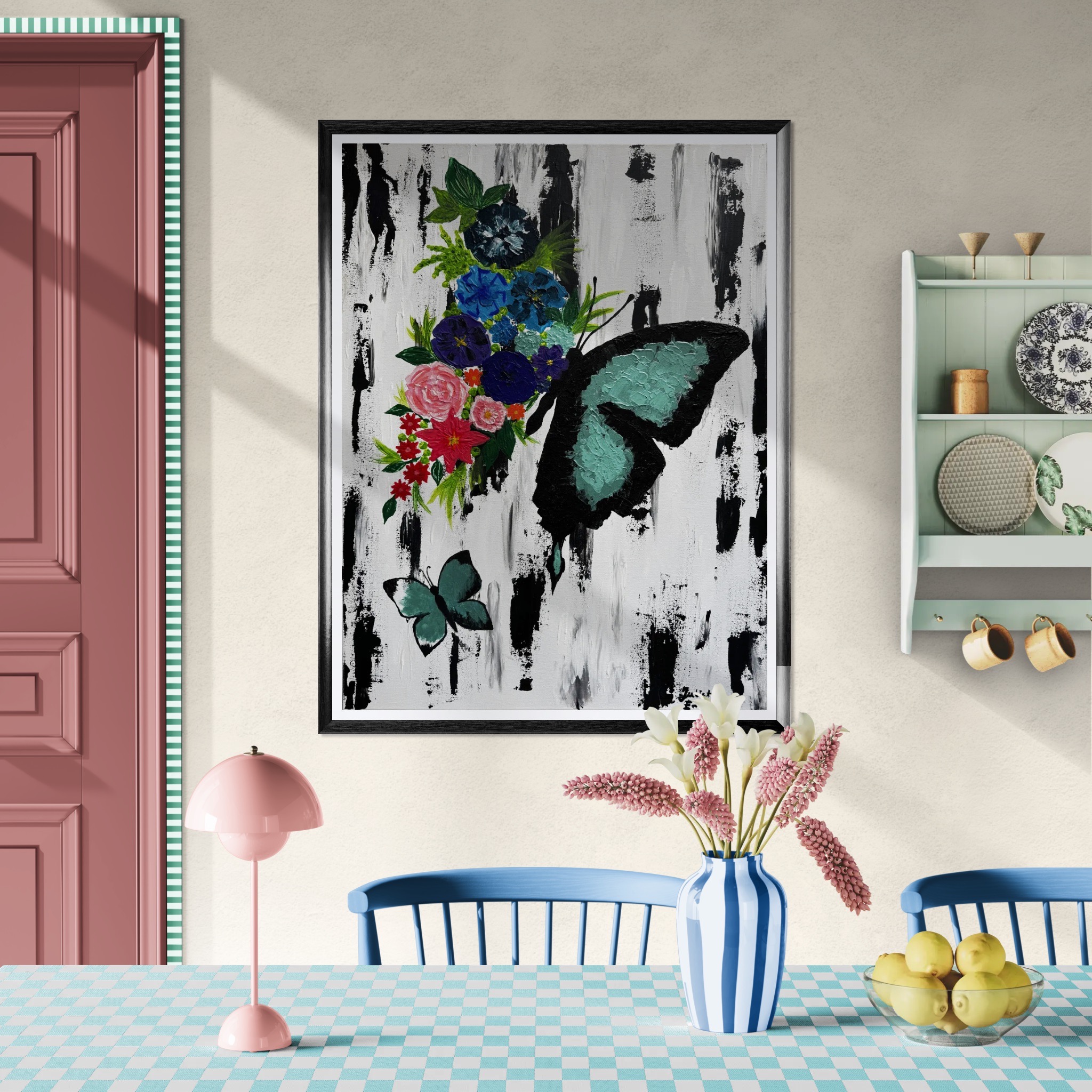 Obraz z motywem motyla i kwiatów w czarnej ramie, wiszący na ścianie. W tle widoczna pastelowa aranżacja wnętrza z elementami wyposażenia.