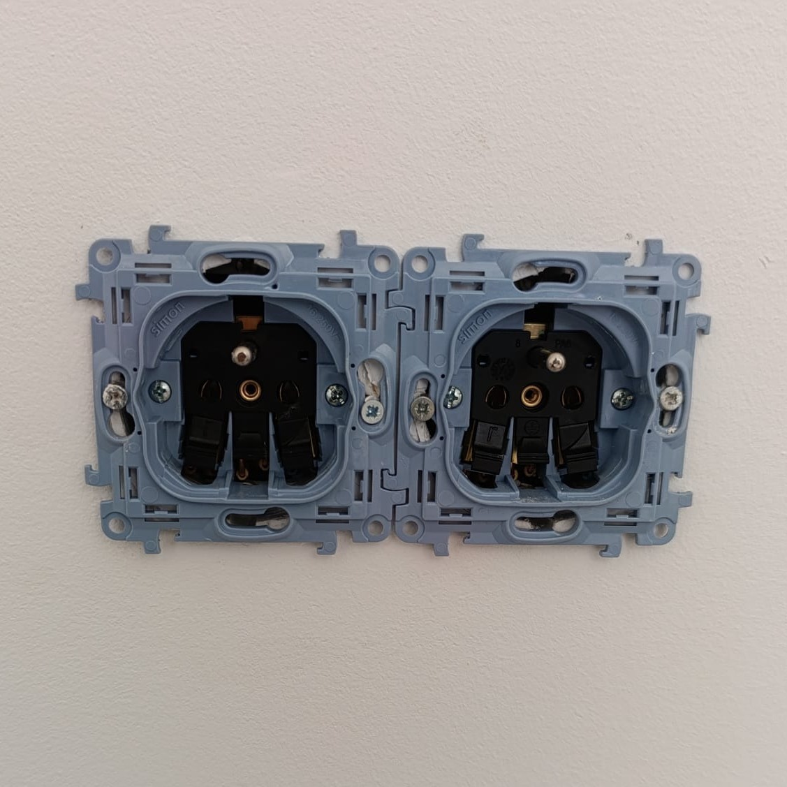 Błękitna, podwójna puszka elektryczna marki Simon osadzona w białej ścianie, gotowa do montażu gniazd. Widoczne śruby mocujące i wnętrze z elementami elektrycznymi.