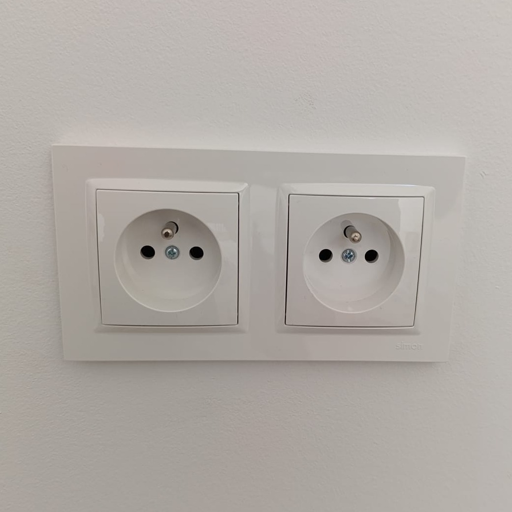 Białe, podwójne gniazdko elektryczne marki Simon, zamontowane na białej ścianie. Widoczne detale montażu i symetryczny układ.