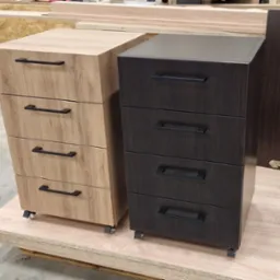 Dwie szafki z szufladami na k&oacute;łkach: jasna drewnopodobna i ciemna, obie z czarnymi uchwytami. Widoczne materiały stolarskie w tle, sugerujące warsztat.