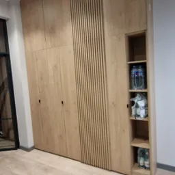 Duża, jasna szafa wnękowa z drewnianymi lamelami i otwartymi półkami, wypełnionymi butelkami wody. Podłoga z jasnego drewna. Nowoczesny design.