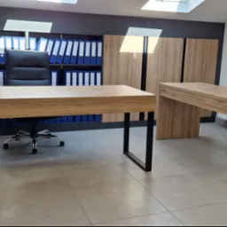 Biuro z dwoma biurkami z drewnianym blatem i czarnymi nogami. W tle regał z segregatorami i szafki w tym samym stylu co biurka. Krzesło biurowe.