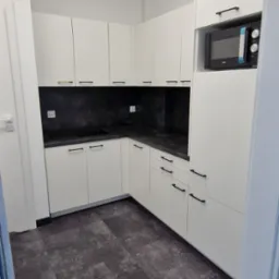 Jasna kuchnia na wymiar z czarnym blatem i wbudowaną mikrofal&oacute;wką. Minimalistyczny design, nowoczesne wykończenie, szare panele podłogowe.