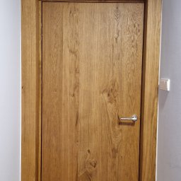 Adam The Partner ADAM DĄBROWSKI - Drewniane drzwi z ościeżnicą, dopasowane kolorystycznie do podłogi. Widoczna faktura drewna i chromowana klamka. Montaż w wąskim korytarzu.