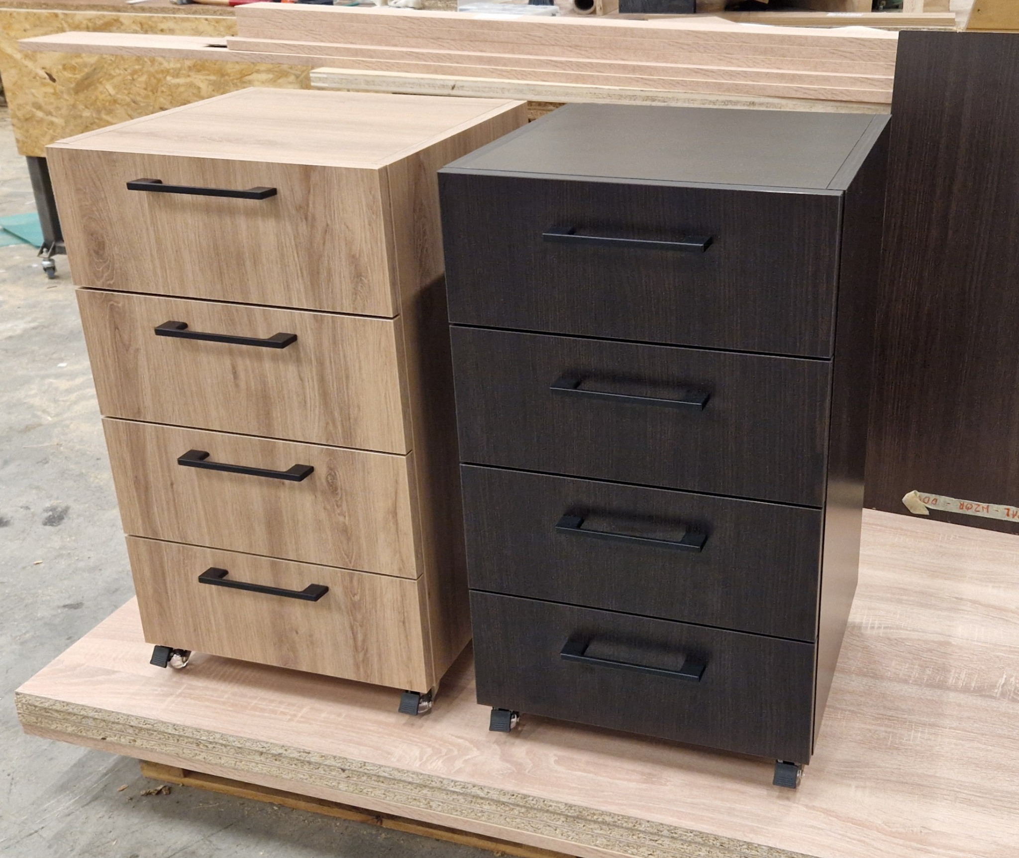 Dwie szafki z szufladami na kółkach: jasna drewnopodobna i ciemna, obie z czarnymi uchwytami. Widoczne materiały stolarskie w tle, sugerujące warsztat.