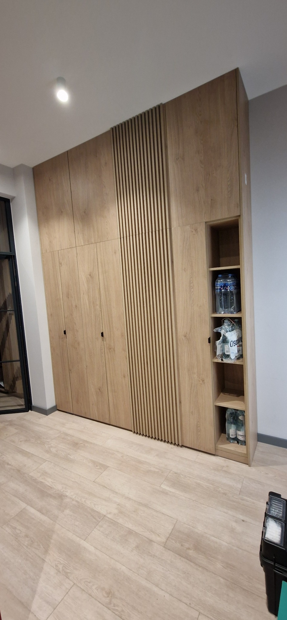 Duża, jasna szafa wnękowa z drewnianymi lamelami i otwartymi półkami, wypełnionymi butelkami wody. Podłoga z jasnego drewna. Nowoczesny design.