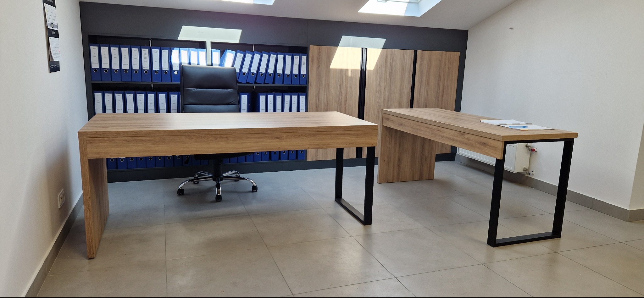 Biuro z dwoma biurkami z drewnianym blatem i czarnymi nogami. W tle regał z segregatorami i szafki w tym samym stylu co biurka. Krzesło biurowe.