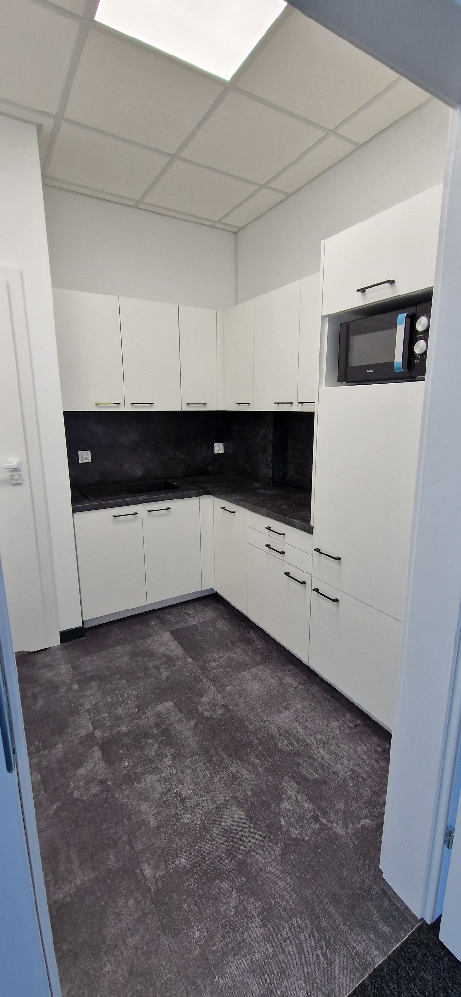 Jasna kuchnia na wymiar z czarnym blatem i wbudowaną mikrofalówką. Minimalistyczny design, nowoczesne wykończenie, szare panele podłogowe.