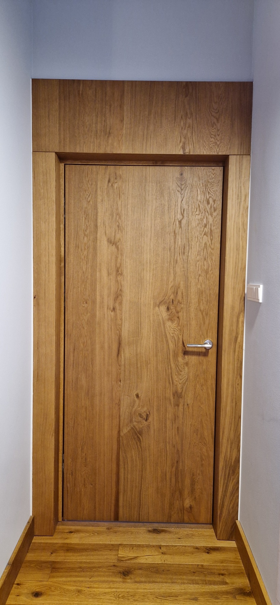 Drewniane drzwi z ościeżnicą, dopasowane kolorystycznie do podłogi. Widoczna faktura drewna i chromowana klamka. Montaż w wąskim korytarzu.