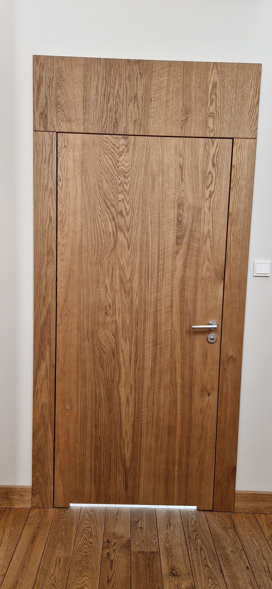 Drewniane drzwi wewnętrzne z ukrytą ościeżnicą, dopasowane do podłogi, z widocznym przełącznikiem światła na ścianie.