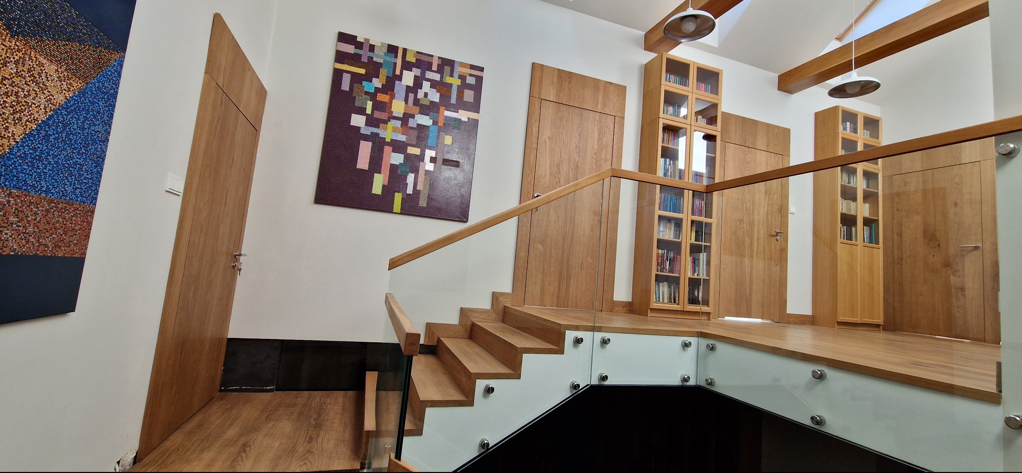 Nowoczesne schody z drewnianymi stopniami i szklaną balustradą. W tle zabudowa z drewnianych szaf i drzwi, oraz abstrakcyjny obraz na ścianie. Jasne oświetlenie.