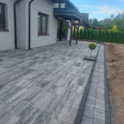 Ułożona kostka brukowa w szaro-białej kolorystyce przed domem z małym drzewkiem wkomponowanym w nawierzchnię. Widoczne obrzeża i fragment rabaty.