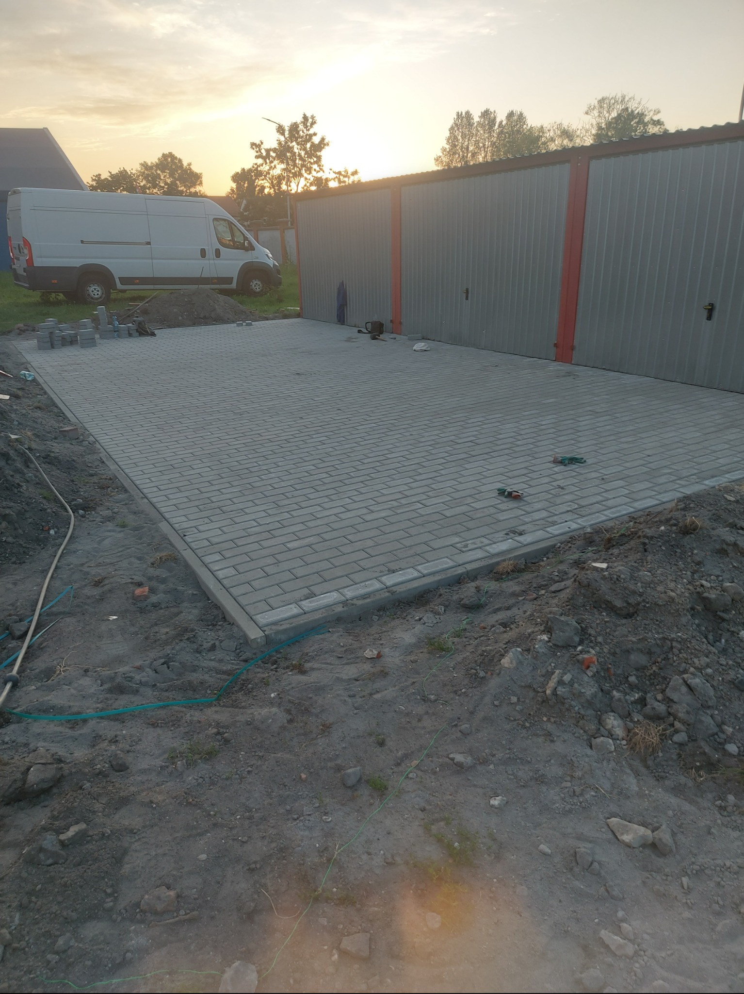 Ułożona kostka brukowa w kształcie prostokąta, obok garaże blaszane i zaparkowany bus. Widoczne narzędzia i resztki materiałów budowlanych. Zachód słońca w tle.