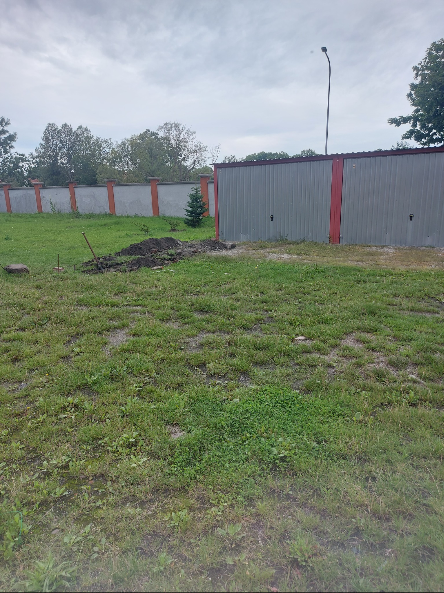 Teren z wykopaną ziemią, narzędzia wbite w ziemię, widoczny garaż blaszany i fragment ogrodzenia. Przygotowanie terenu pod kostkę lub niwelacja.