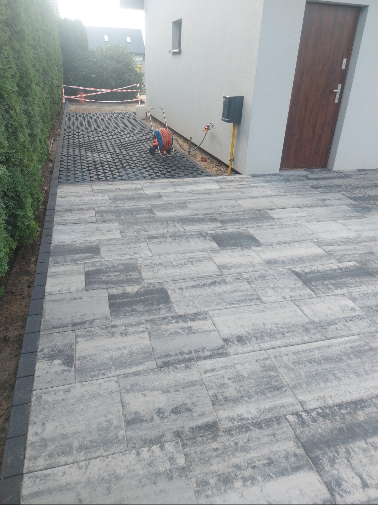 Ułożona szara kostka brukowa z obrzeżem, widoczne także czarne płyty ażurowe. W tle budynek i zwinięty wąż ogrodowy. Całość w naturalnym oświetleniu.