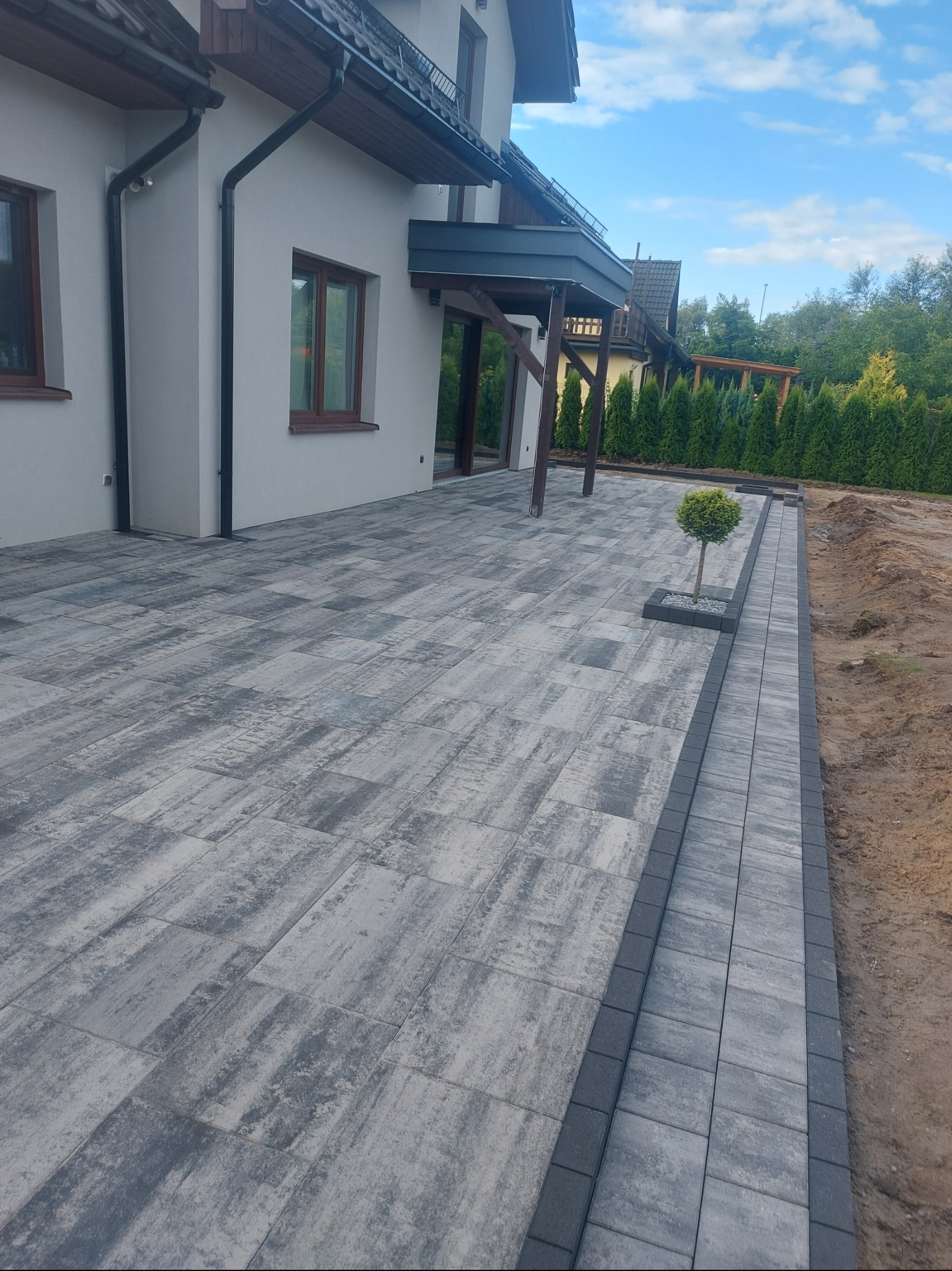 Ułożona kostka brukowa w szaro-białej kolorystyce przed domem z małym drzewkiem wkomponowanym w nawierzchnię. Widoczne obrzeża i fragment rabaty.