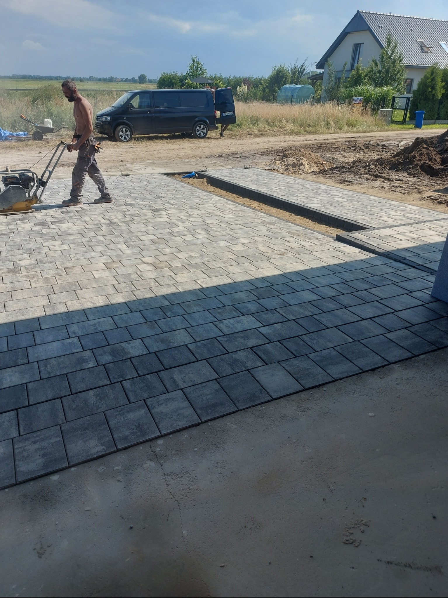 Mężczyzna ubija kostkę brukową szarą zagęszczarką na posesji. Częściowo ułożona nawierzchnia z kostki, w tle dom i samochód dostawczy.