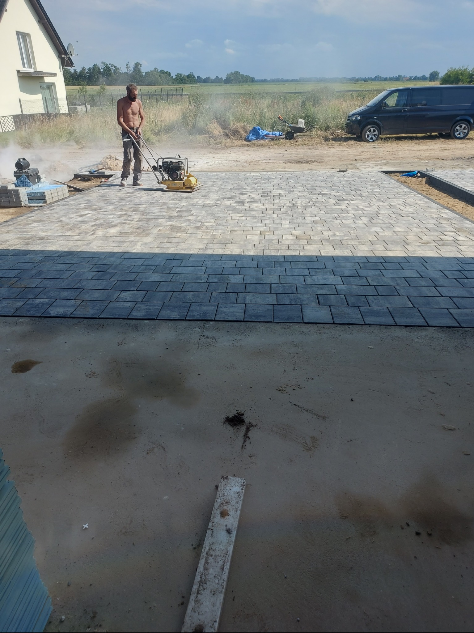 Pracownik zagęszcza wibroprasą ułożoną, szarą kostkę brukową na posesji. Częściowo widoczny dom, samochód i narzędzia.