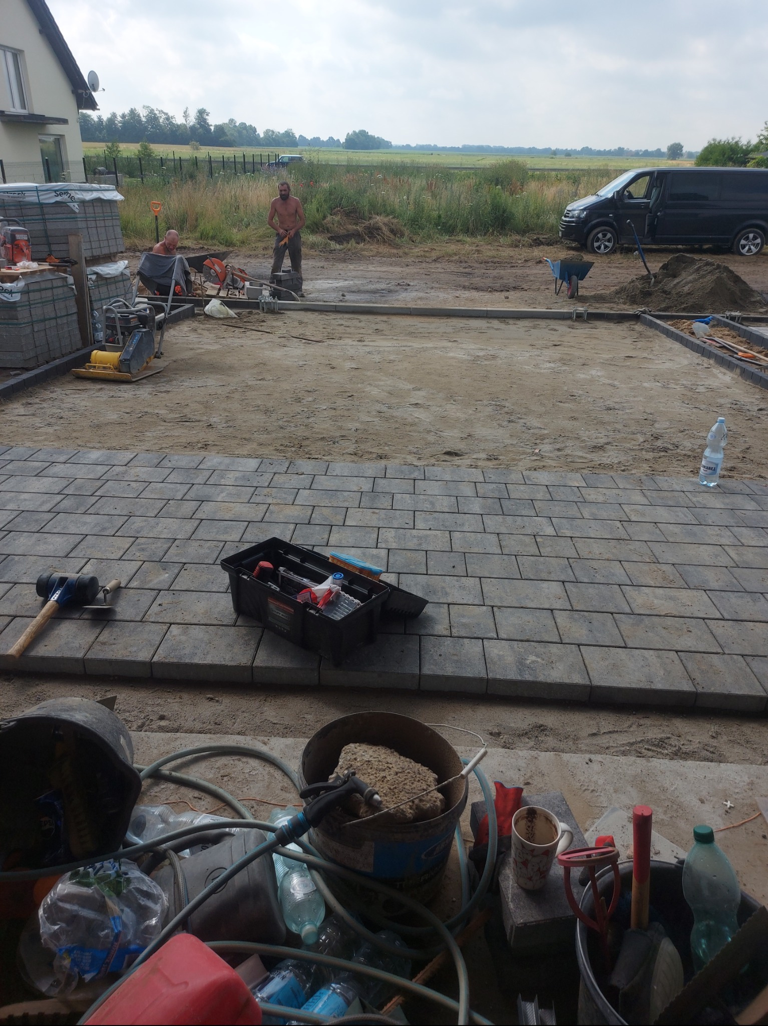 Etap układania szarej kostki brukowej na posesji. Widoczne narzędzia, materiały budowlane i dwóch pracowników w trakcie pracy. W tle krajobraz wiejski i czarny van.