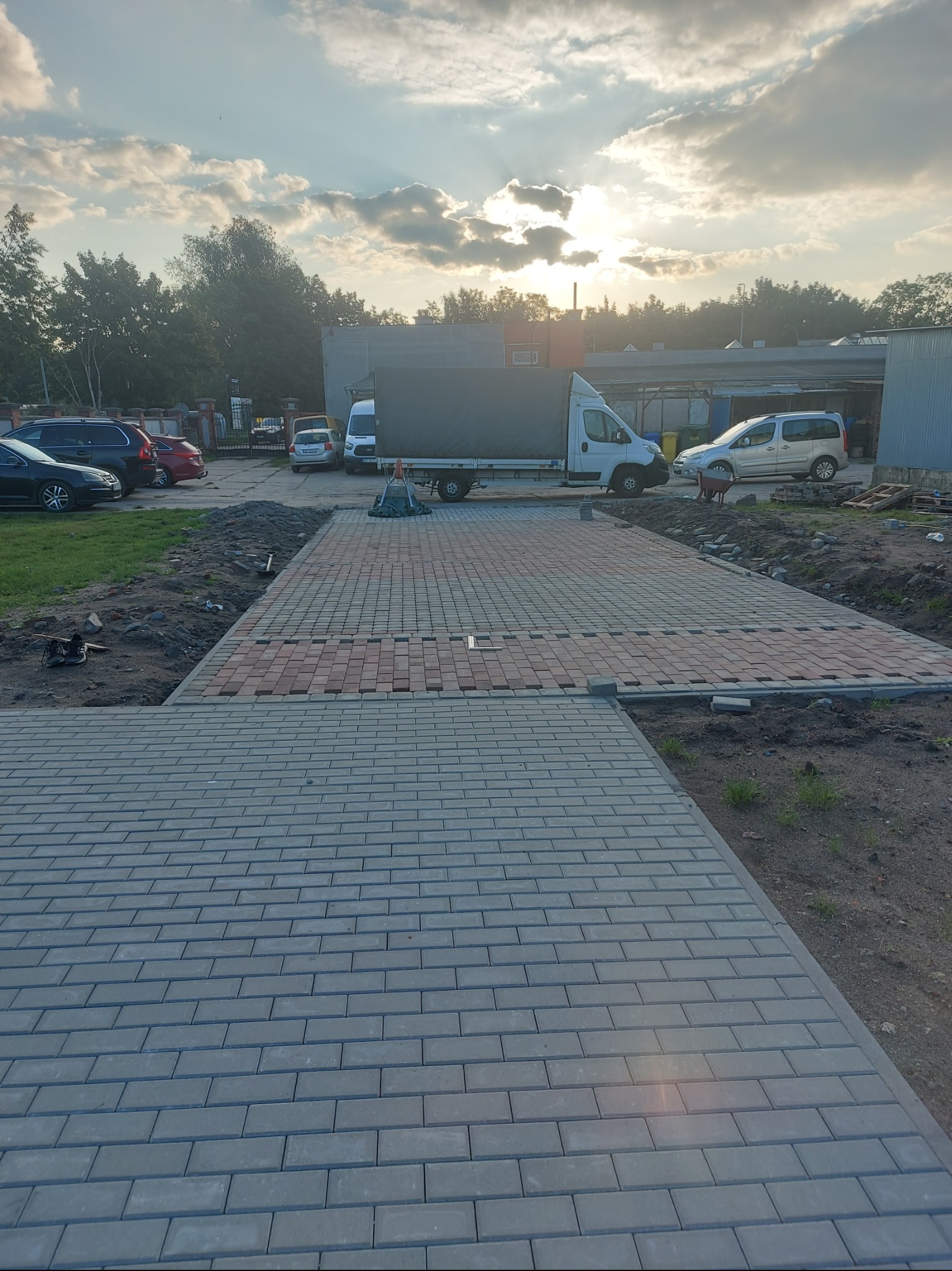 Fragment ułożonej kostki brukowej w dwóch kolorach, z pojazdami w tle. Widoczne ślady prac ziemnych. Słońce przebija się przez chmury.