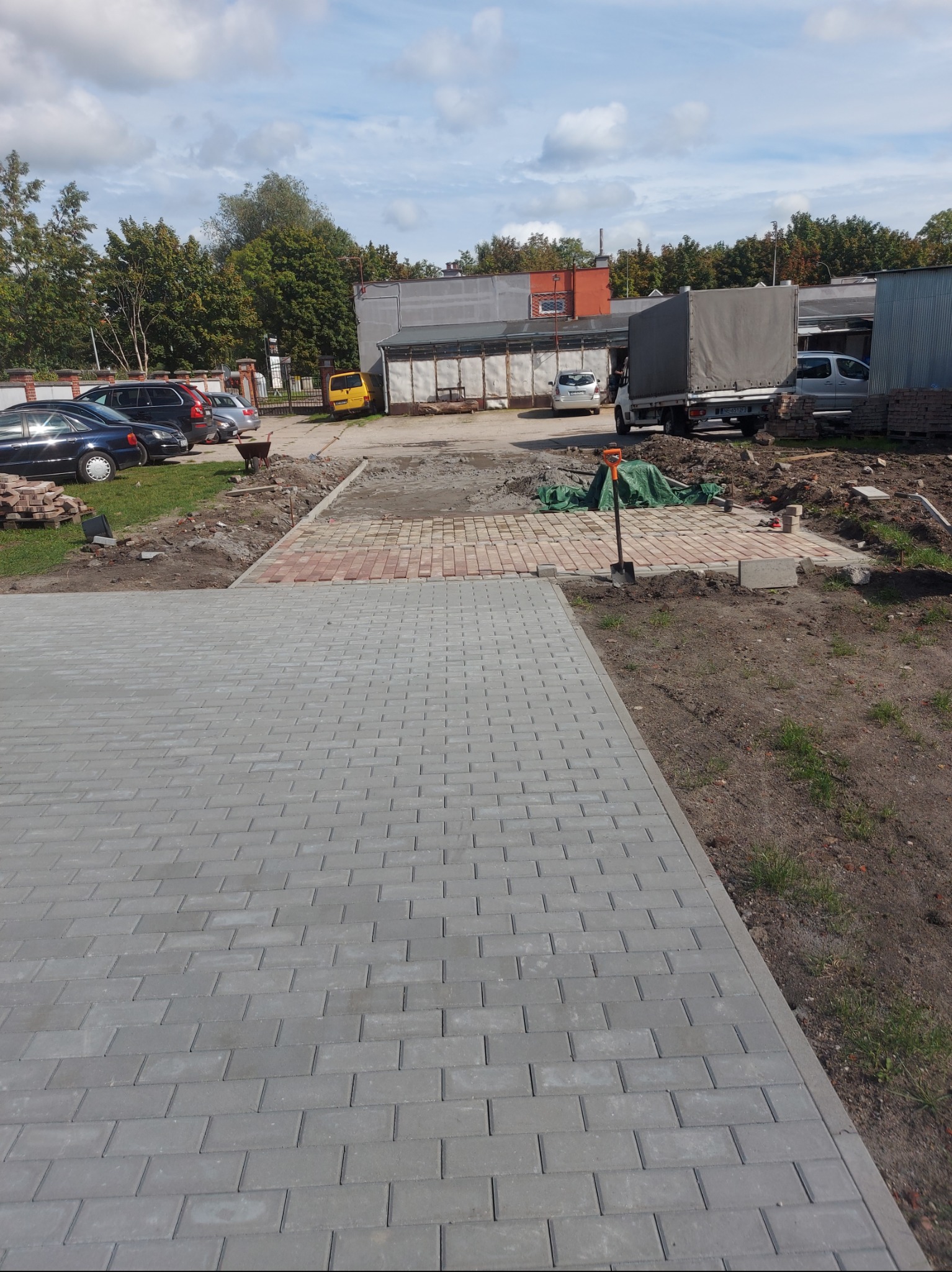 Fragment nowej nawierzchni z szarej kostki, dalsza część w trakcie układania z czerwonej kostki, w tle zaparkowane auta i budynki gospodarcze.