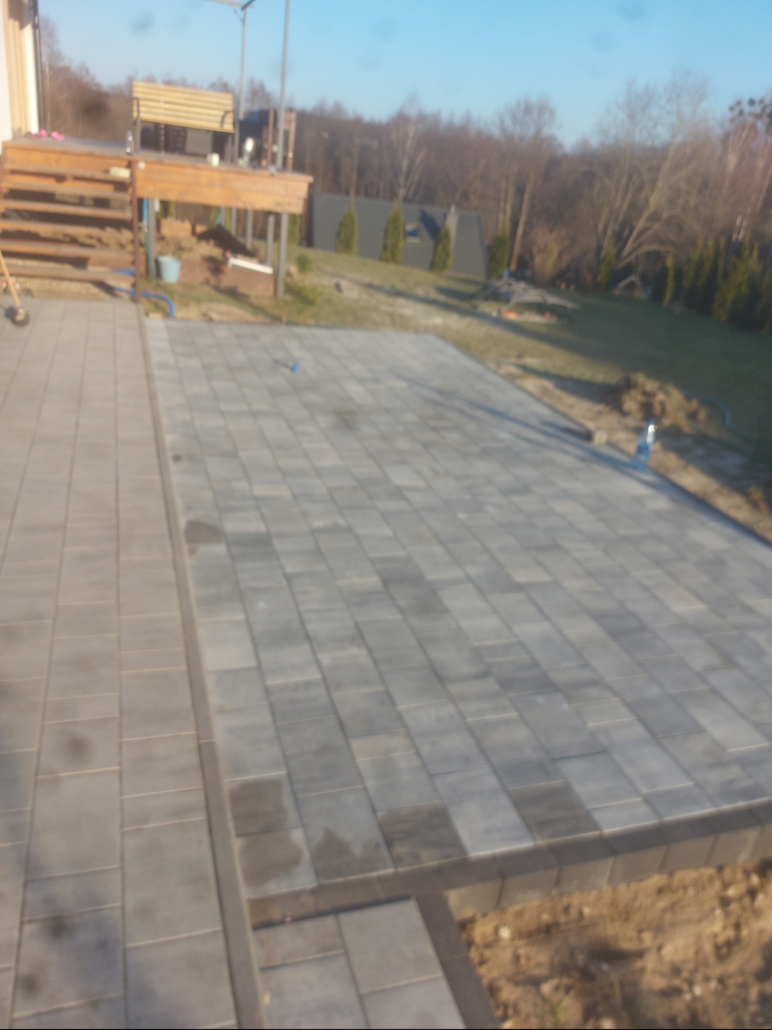 Ułożona, szara kostka brukowa na posesji z drewnianym tarasem i zielenią w tle. Widoczne detale wykończenia i kształt kostki.